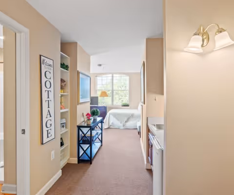 Interior view of a senior living facility room at Spring Hills Mount Vernon, showing a hallway leading to a bedroom with a large window. The hallway has a small blue shelf with plants and decorations, a wall sign that says 'Welcome to the Cottage,' and a kitchenette area with a sink and cabinets on the right. The room is well-lit with beige walls and carpeted floor.