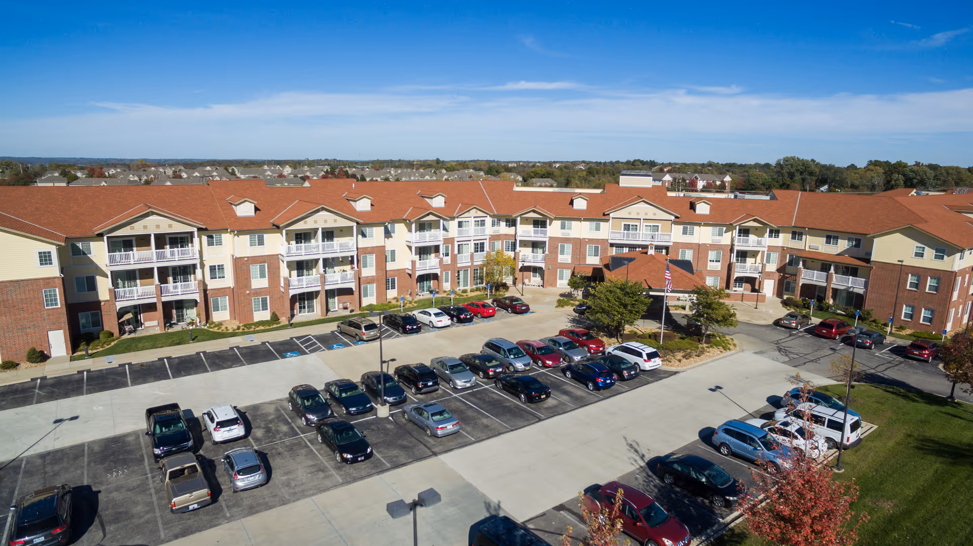 Aerial view of a large senior living facility named Shawnee Hills with a red roof and multiple balconies. The building surrounds a parking lot filled with cars. The sky is clear and blue, and there are trees and greenery around the property.