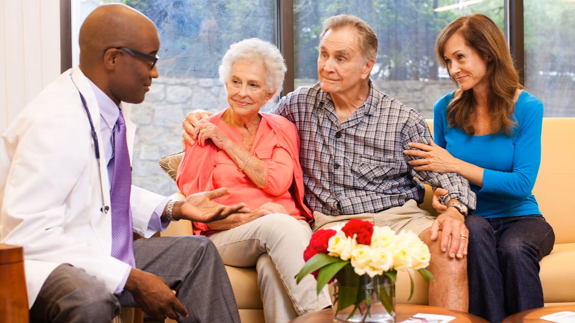 A doctor converses with an elderly couple and a woman in a comfortable seating area, with a flower arrangement in front.