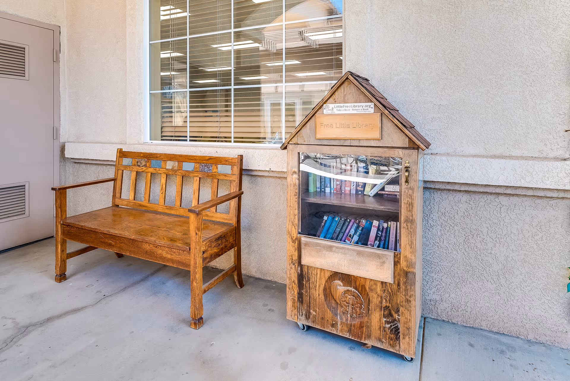 A wooden bench sits beside a small wooden free little-library box against the exterior wall of a senior living building.