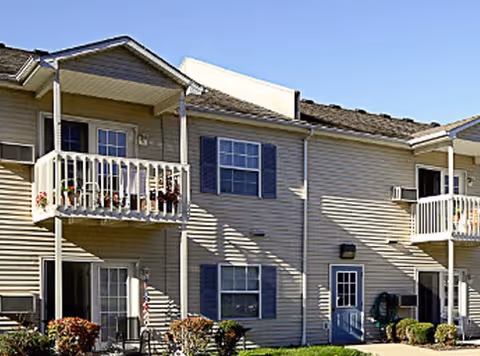 Two-story beige senior apartment building with balconies, blue shutters, and a small front lawn.