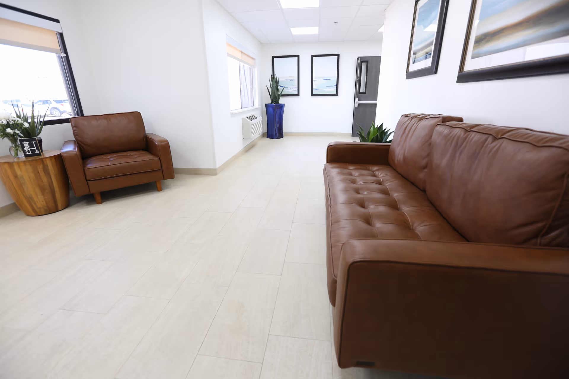 Bright waiting room with a brown leather sofa and armchair, potted plants, framed art, and a light tiled floor.