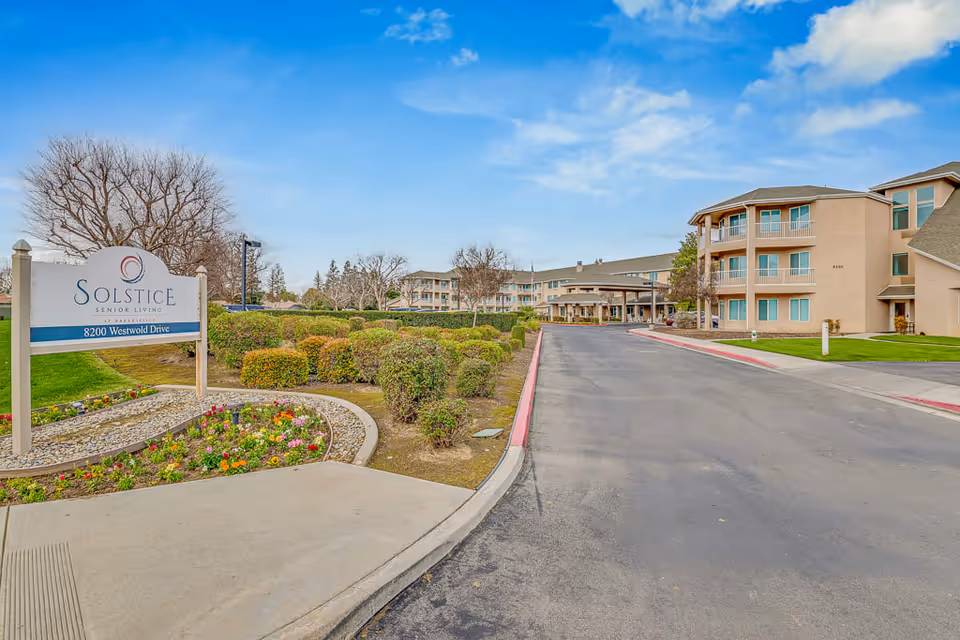 Exterior view of Solstice Senior Living at Bakersfield showing the entrance driveway, landscaped bushes, flower bed, and the multi-story beige building under a partly cloudy blue sky.
