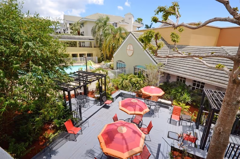 Outdoor patio area with multiple red and orange umbrellas shading tables and chairs, surrounded by greenery and trees. In the background, there is a swimming pool and several buildings with light-colored walls and tiled roofs under a blue sky with some clouds.
