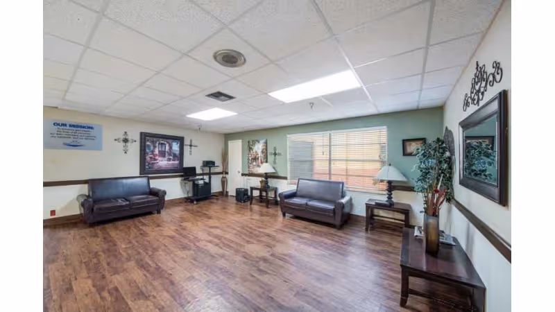 A spacious room with wooden flooring and a drop ceiling, furnished with two dark brown leather sofas, two side tables with lamps, a console table with decorative items, and wall art including crosses and framed pictures. A window with blinds lets in natural light.