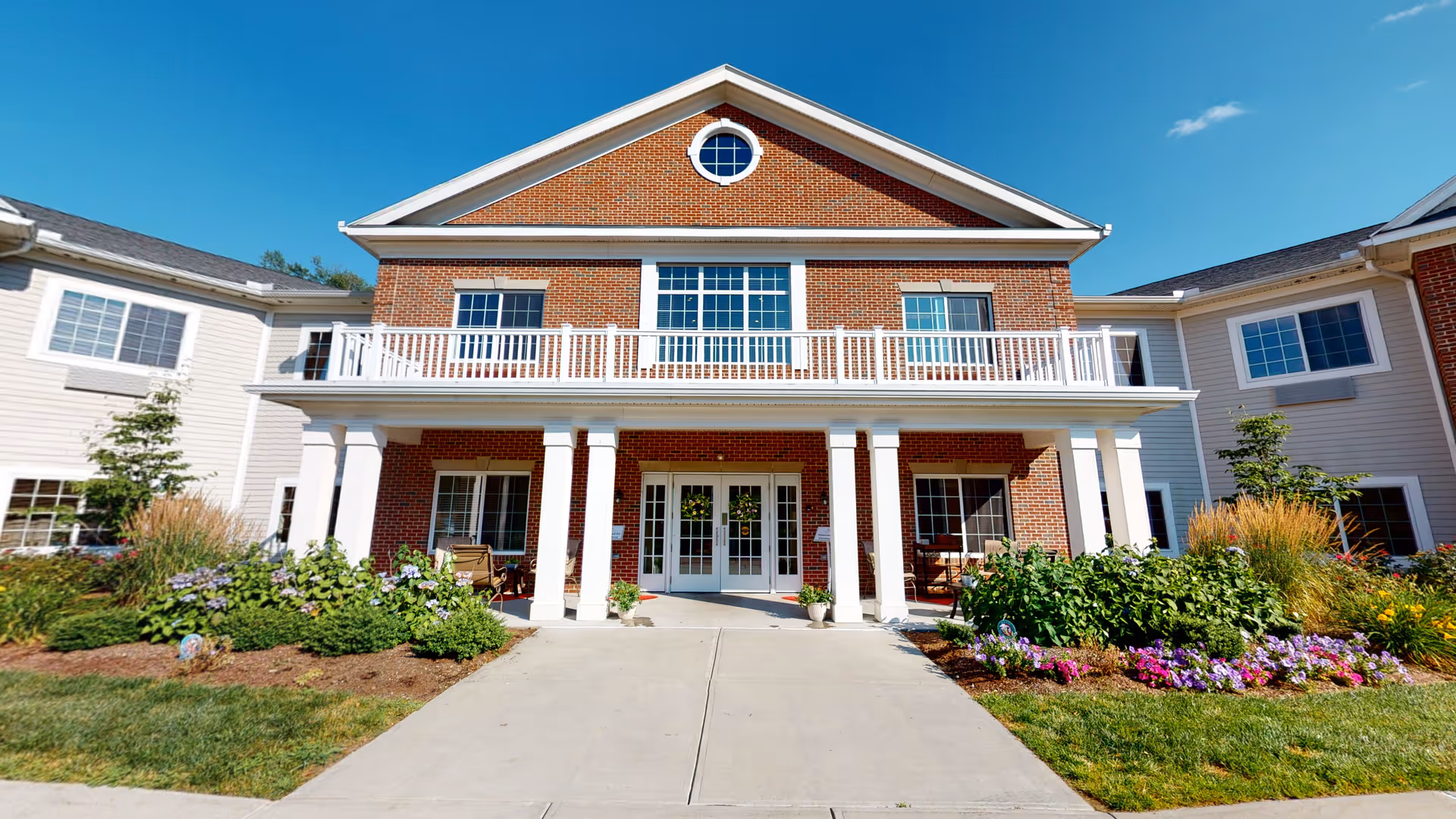 Front exterior view of a two-story senior living facility building with a brick facade in the center and white siding on the wings. The entrance features white columns supporting a balcony with white railing. There are landscaped flower beds with green shrubs and colorful flowers on either side of the concrete walkway leading to the entrance. The sky is clear and blue.
