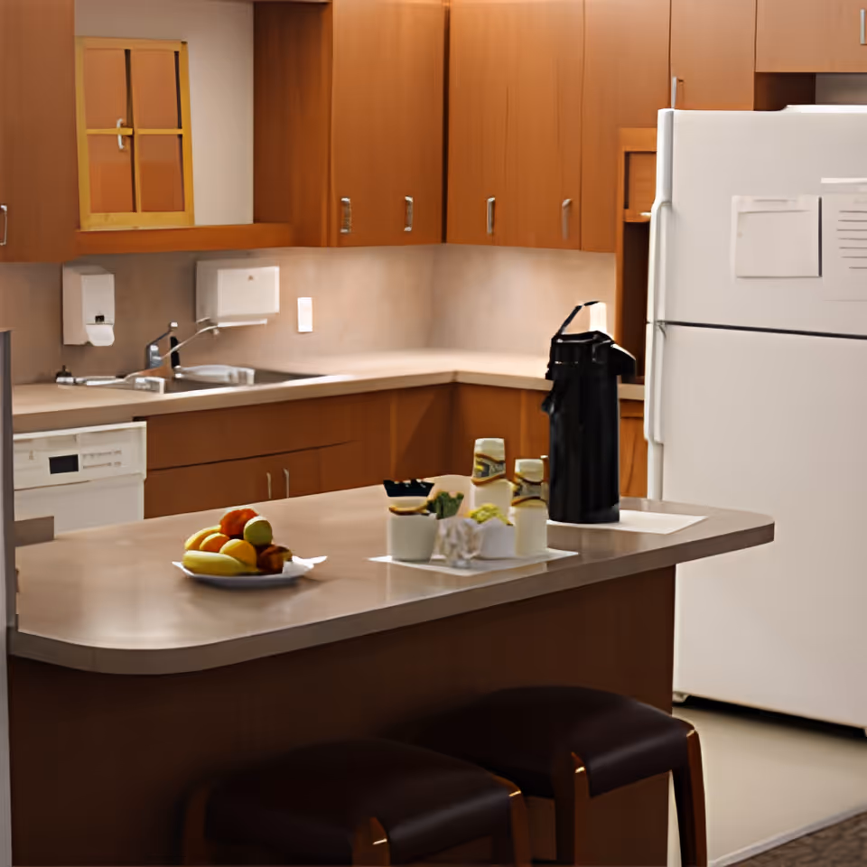 A kitchen area with wooden cabinets, a white refrigerator, a dishwasher, and a countertop island with two stools. On the countertop, there is a plate of assorted fruits, a black thermos, and several condiment containers.