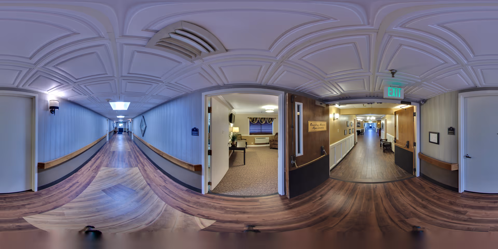Wide-angle view of a long, well-lit hallway in a senior living facility with wood flooring and light blue walls. On the right side, there is an open door leading to a room with carpeted floor, a window with curtains, and some furniture. Further down the hallway, there is another corridor visible with additional doors and seating. The ceiling has a decorative pattern and there is an exit sign above one of the doors.