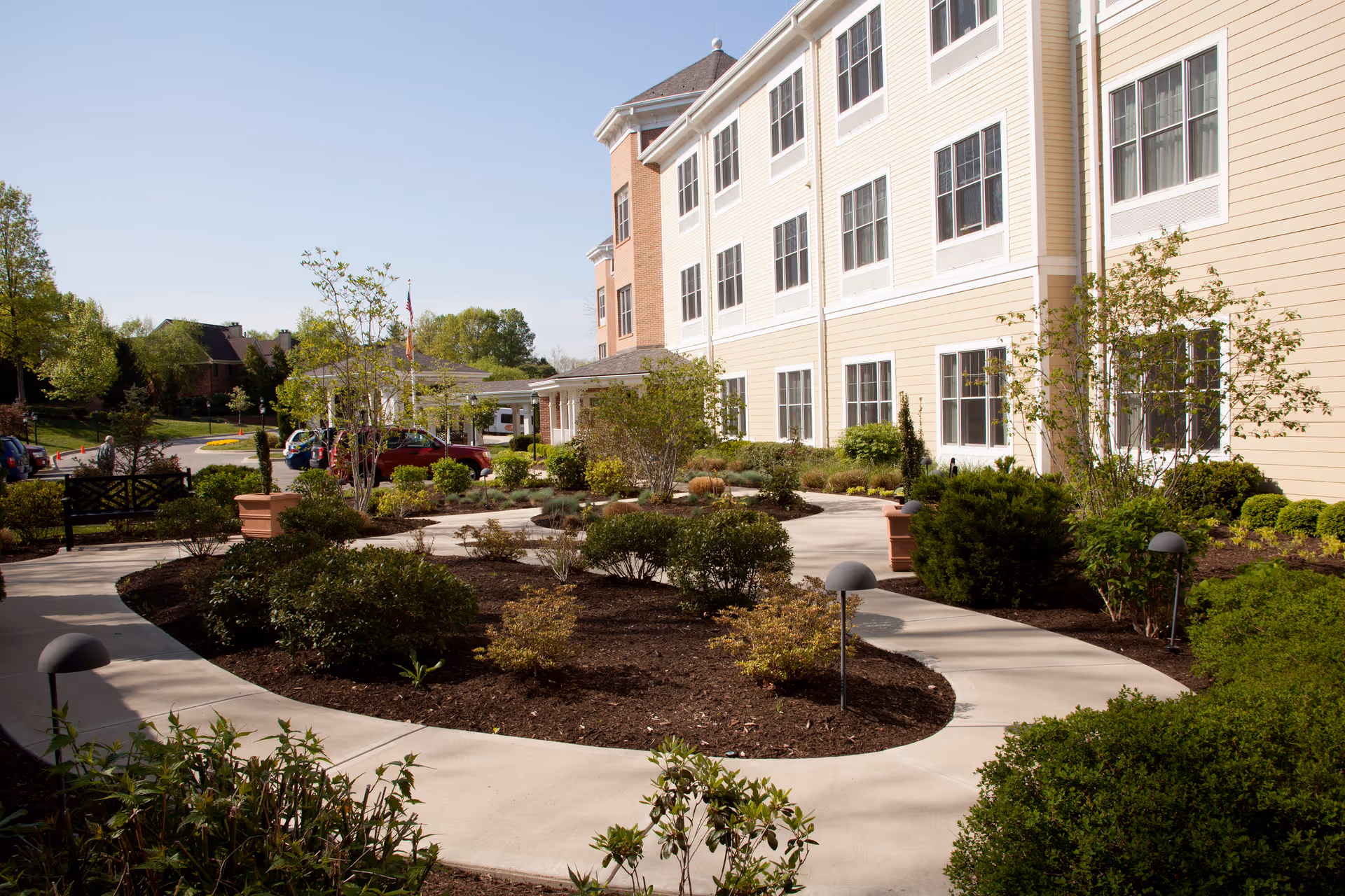 Landscaped courtyard with a curved sidewalk in front of a multi-story beige senior living building.