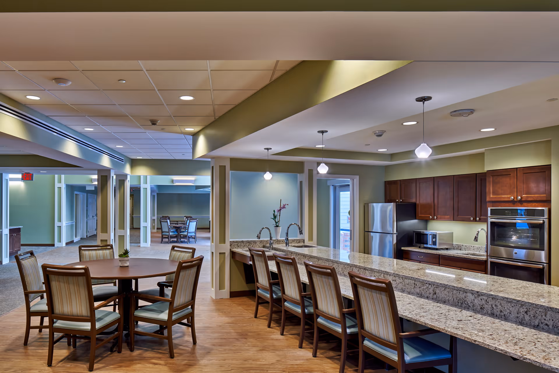 A spacious and modern kitchen and dining area in a senior living facility. The kitchen features granite countertops, wooden cabinets, a stainless steel refrigerator, microwave, and built-in oven. There is a long counter with several chairs for seating. Adjacent to the kitchen is a round dining table with six chairs. The room has soft green walls, wood flooring, and recessed lighting with pendant lights above the counter.