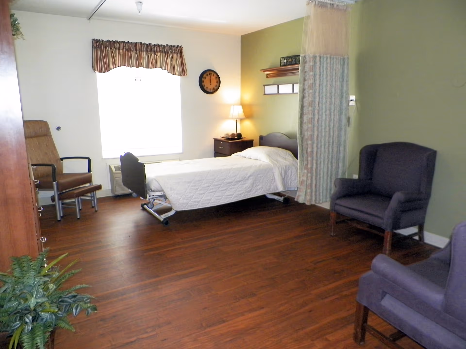 A senior living facility bedroom with a single hospital-style bed covered with a white quilt, a wooden nightstand with a lamp, two upholstered armchairs, a brown leather chair, a window with a striped valance, a wall clock, and a potted plant on the wooden floor.