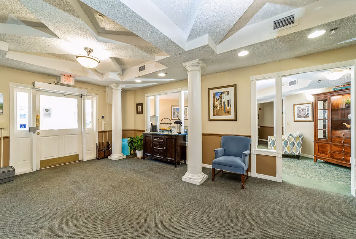Interior view of a senior living facility lobby area with beige walls and carpeted floor. There are two white decorative columns, a blue armchair, a wooden cabinet with water dispensers and cups, framed artwork on the walls, and a glass door exit with an illuminated exit sign above it. Adjacent room visible with patterned carpet and wooden furniture.