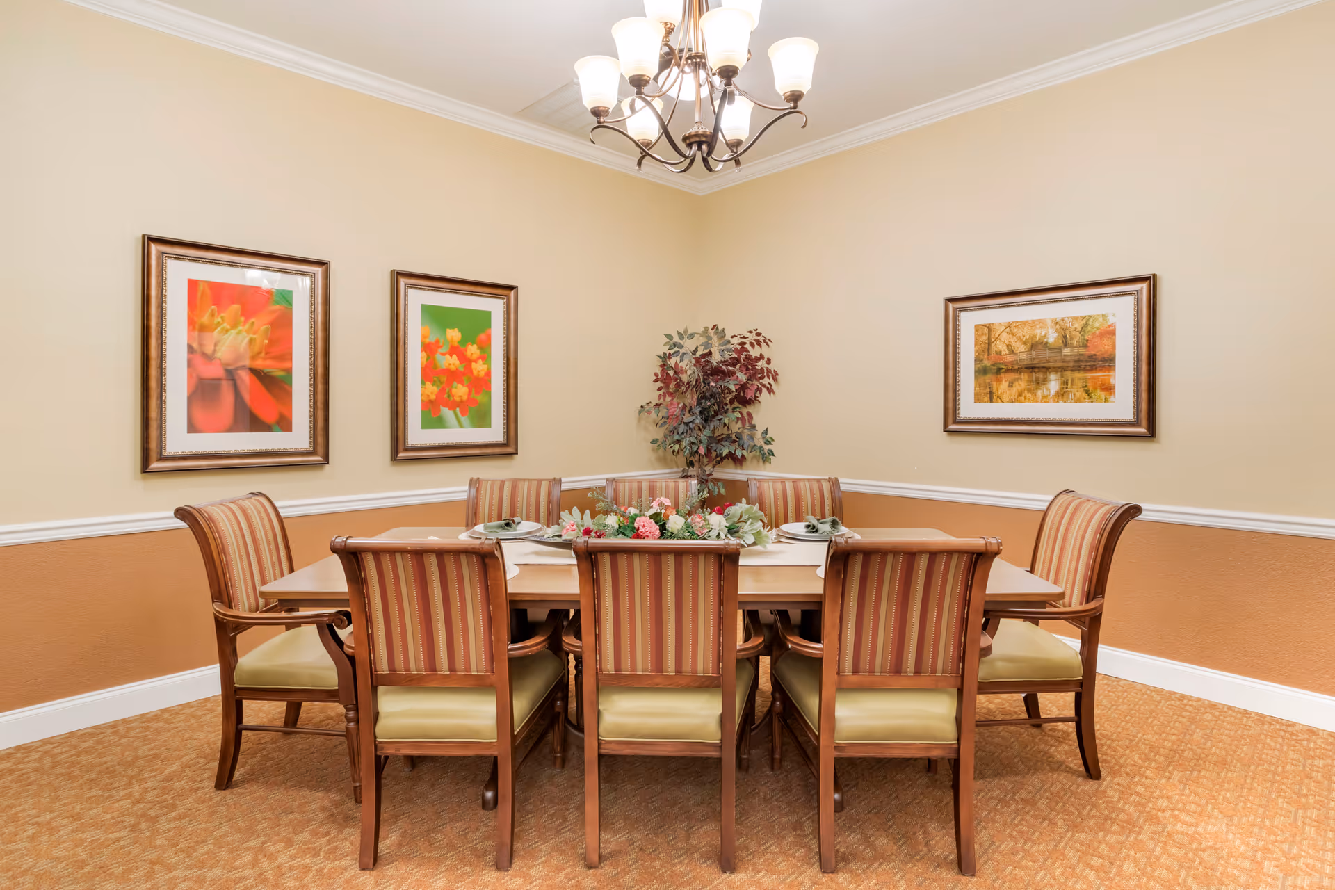 A dining room with a rectangular wooden table surrounded by eight striped upholstered chairs. The table is decorated with a floral centerpiece and place settings. The walls are painted beige with a white chair rail and three framed pictures hanging on the walls. A chandelier with multiple lights hangs from the ceiling, and there is a potted plant in the corner.