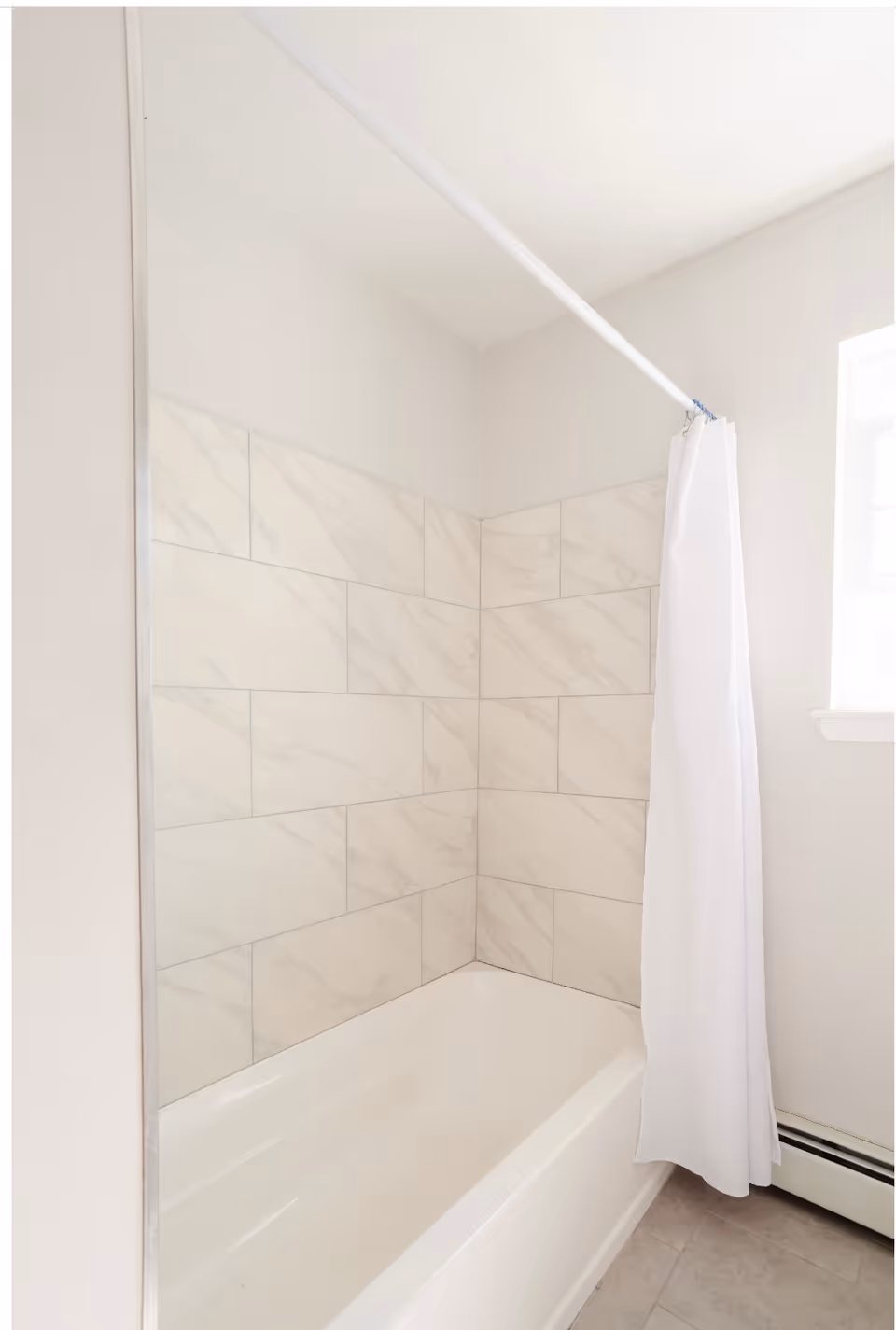 White bathtub with marble-look tiled shower surround and a white shower curtain hanging from a rod near a small window.