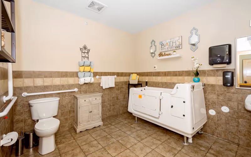 A spacious assisted living bathroom with beige and brown tiled walls and floor. The room features a white toilet with grab bars, a white accessible bathtub with a door, a small wooden cabinet, a towel rack with neatly folded towels in blue, yellow, and white, a wall-mounted paper towel dispenser, a soap dispenser, a mirror, and decorative wall art including a sign that reads 'BATHS with bubbles and relax'.