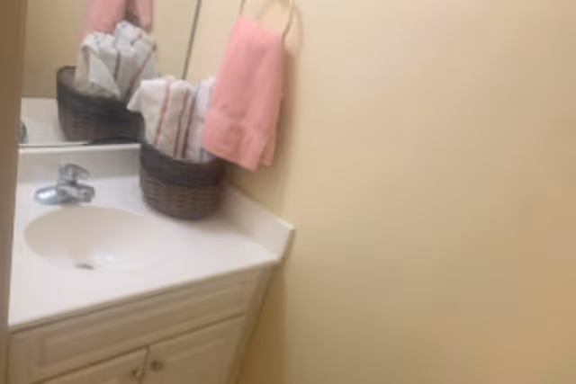A bathroom sink with a white countertop and cabinet below. On the countertop, there is a brown wicker basket holding neatly folded white towels with red stripes. A pink hand towel hangs on a gold towel ring mounted on the beige wall. A mirror is visible behind the sink and basket.