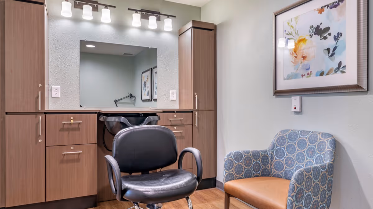 Interior view of a salon area in a senior living facility featuring a black salon chair in front of a sink and mirror, surrounded by wooden cabinets. To the right, there is a patterned armchair with a brown seat cushion and a framed floral artwork on the wall above it.