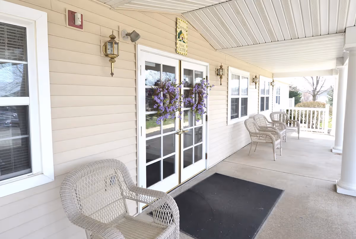 Covered porch area with beige siding, white-framed windows, and a set of double glass doors decorated with purple floral wreaths. Several wicker chairs are arranged along the porch, which has white columns and a concrete floor with a black mat near the doors.