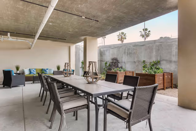 Covered outdoor patio area with a long dining table surrounded by wicker chairs, two decorative lanterns on the table, a seating area with a sofa and armchairs with cushions, and planter boxes with greenery against a concrete wall.