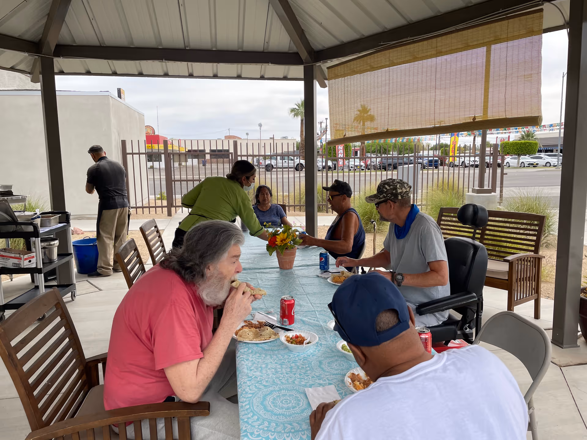 Several people sit and eat around a table under a covered outdoor pavilion while a staff member serves food.