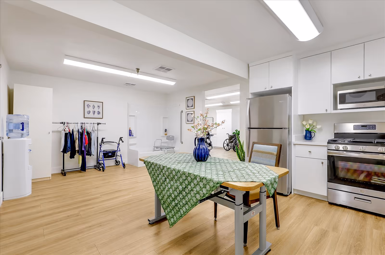 A bright, clean room with a kitchen area featuring a stainless steel refrigerator, stove, and microwave. A table with a green patterned tablecloth and a blue vase with flowers is in the foreground. The room also includes a water cooler, a clothing rack with hanging clothes, a walker, and a wheelchair. The flooring is light wood, and the walls are white with framed artwork.