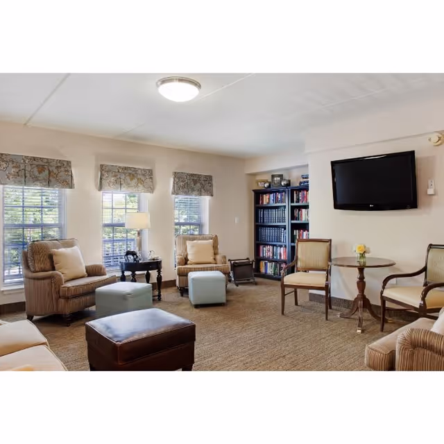 Bright communal living room with armchairs and ottomans arranged around a TV and bookshelf.