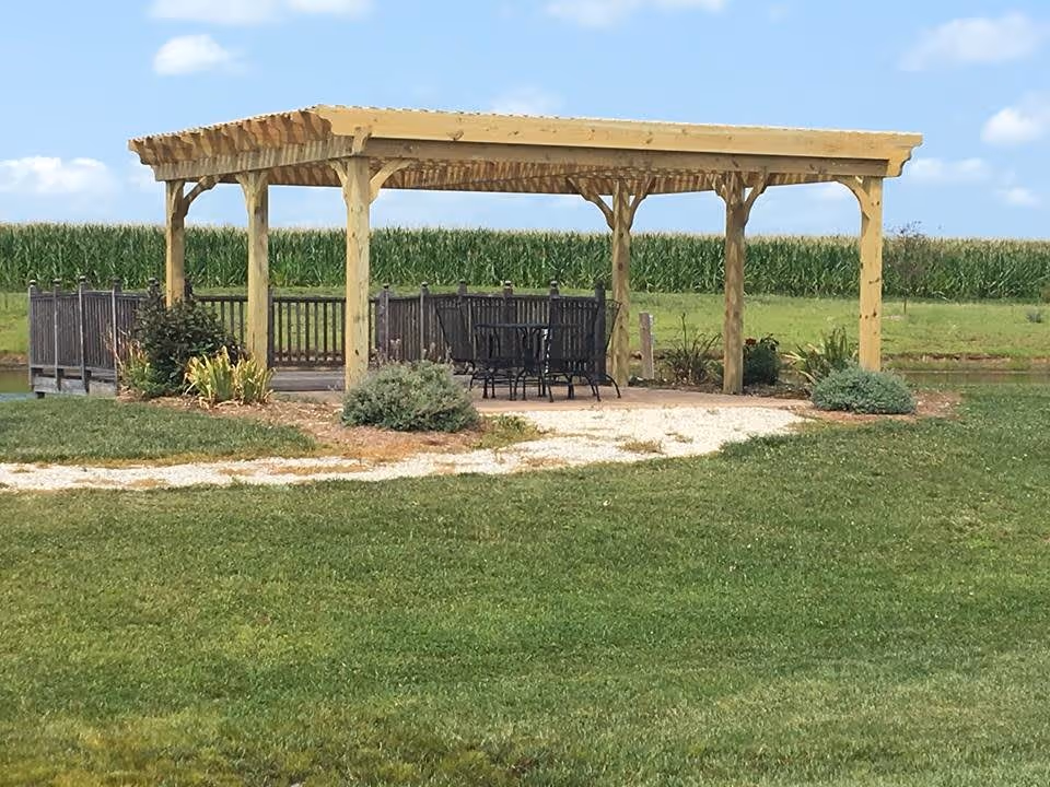 A wooden pergola with a table and several chairs underneath, situated on a grassy area with a gravel path leading to it. In the background, there is a wooden fence and a field of tall green crops under a partly cloudy sky.