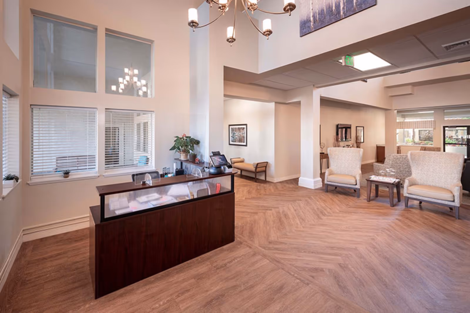 Reception area of Holiday Warburton Place featuring a wooden reception desk with a glass top, a potted plant, and office equipment. The space has light-colored walls, large windows with blinds, wood-patterned flooring, and a seating area with two upholstered armchairs and a small table. A chandelier hangs from the ceiling, and there is artwork on the walls.