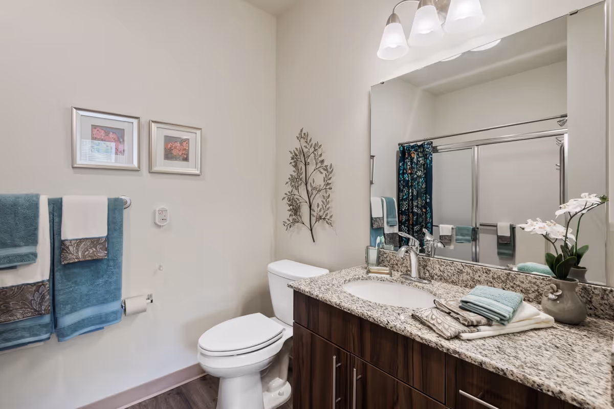 A clean and modern bathroom featuring a white toilet, a granite countertop with a sink, and a large mirror above the sink. The countertop has neatly folded towels and a small vase with white flowers. On the wall above the toilet, there is a decorative metal wall art piece and two framed pictures. A towel rack with teal and white towels is mounted on the wall next to the toilet. In the reflection of the mirror, a shower with sliding glass doors and a floral shower curtain is visible.