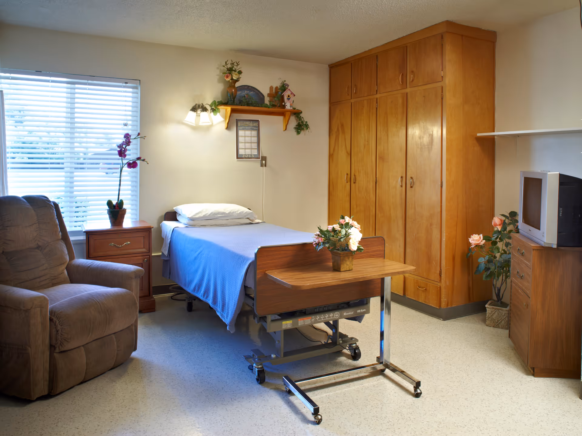 A senior living facility bedroom with a single bed covered in a blue blanket, a brown recliner chair, a wooden nightstand with a potted orchid, a wooden wardrobe, a small TV on a wooden dresser, and a rolling overbed table with a flower arrangement. The room has a window with blinds and a wall-mounted light fixture above the bed.