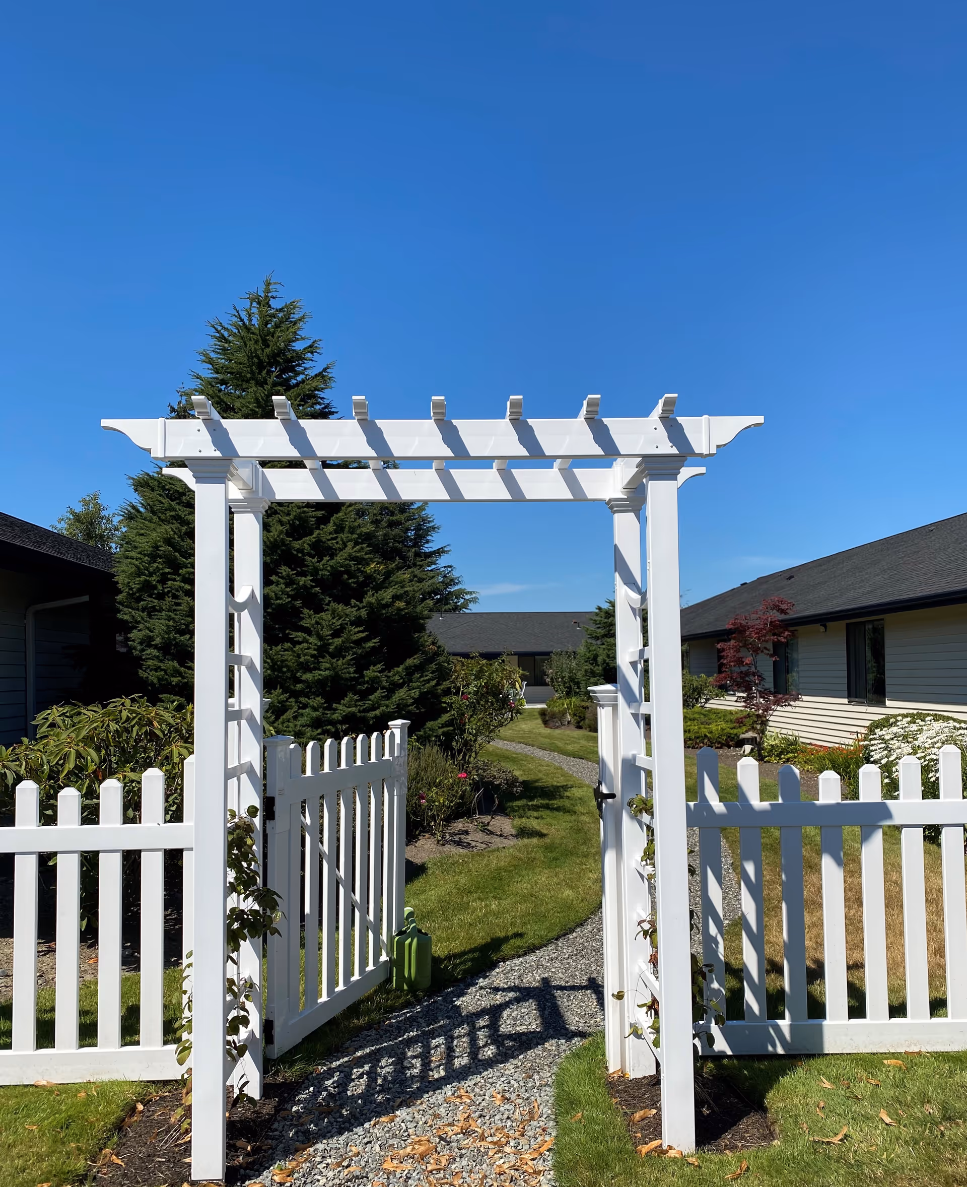 White wooden garden archway with a white picket fence gate open, leading to a gravel path surrounded by green grass, bushes, and trees under a clear blue sky at Logan Creek Retirement Community.
