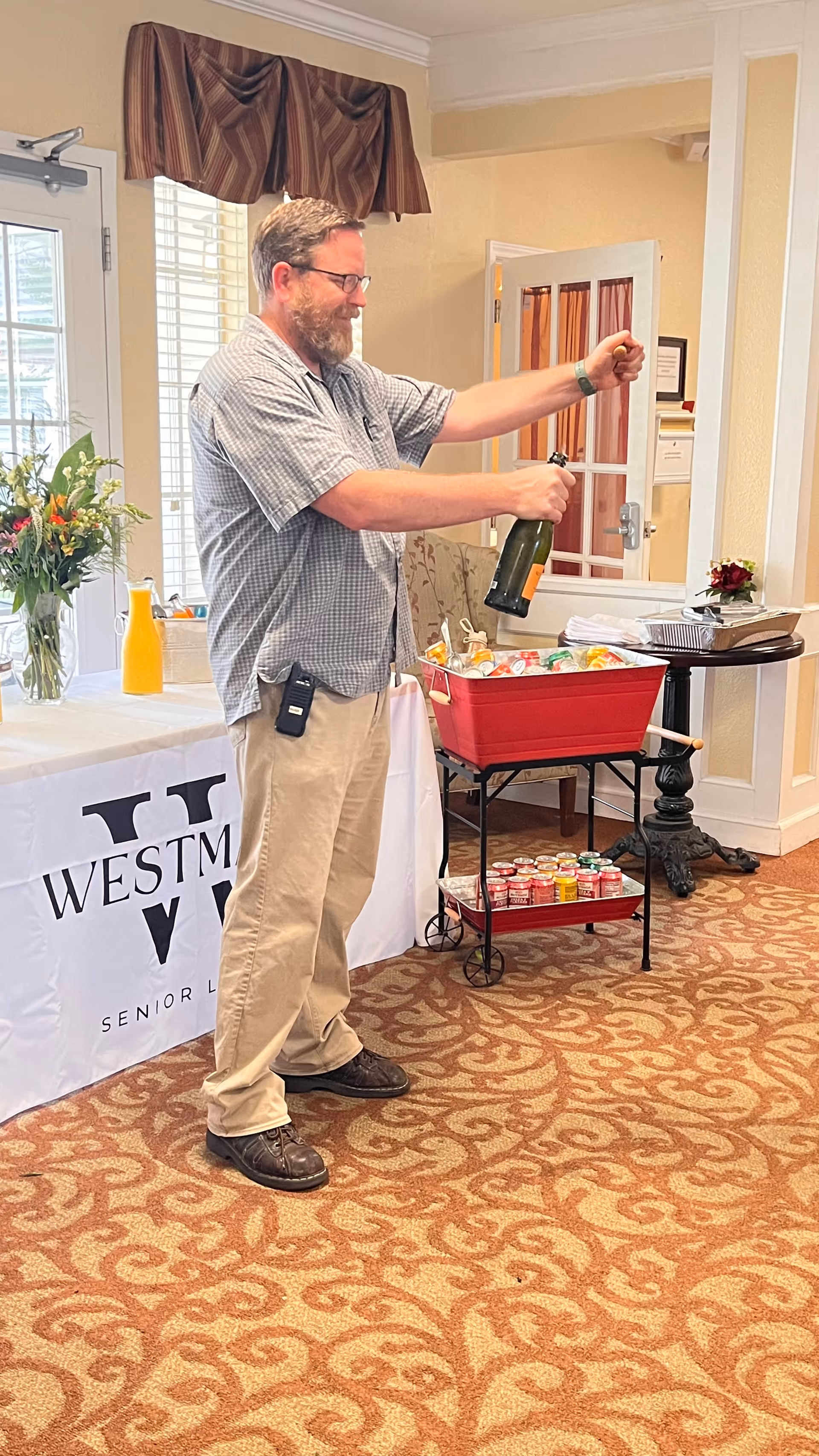 A man with glasses and a beard is opening a bottle of champagne in a room with patterned carpet and light-colored walls. Behind him is a table with a white tablecloth that has the WestMark Senior Living logo, a vase with flowers, and a bottle of orange juice. Next to the table is a red cooler filled with ice and drinks, and a small table with additional items.
