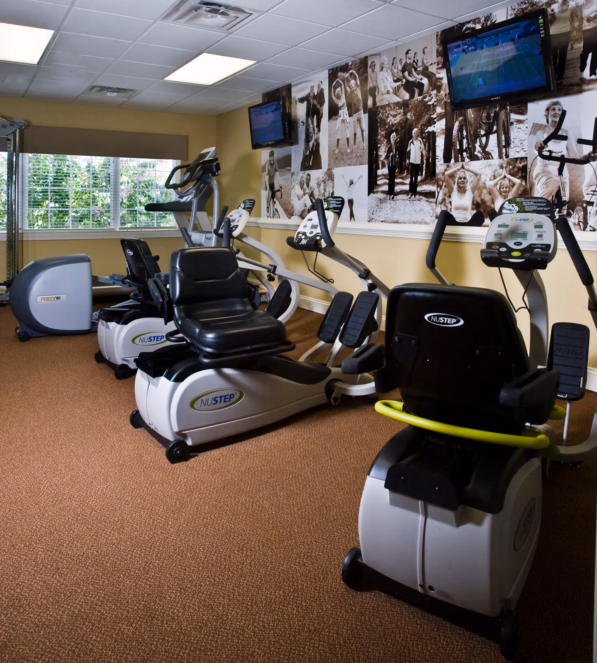 Indoor exercise room with several recumbent stationary bikes and other fitness equipment. The room has carpeted flooring, a window with blinds showing greenery outside, and a wall decorated with black and white photos of people engaging in various physical activities. Two televisions are mounted on the wall.