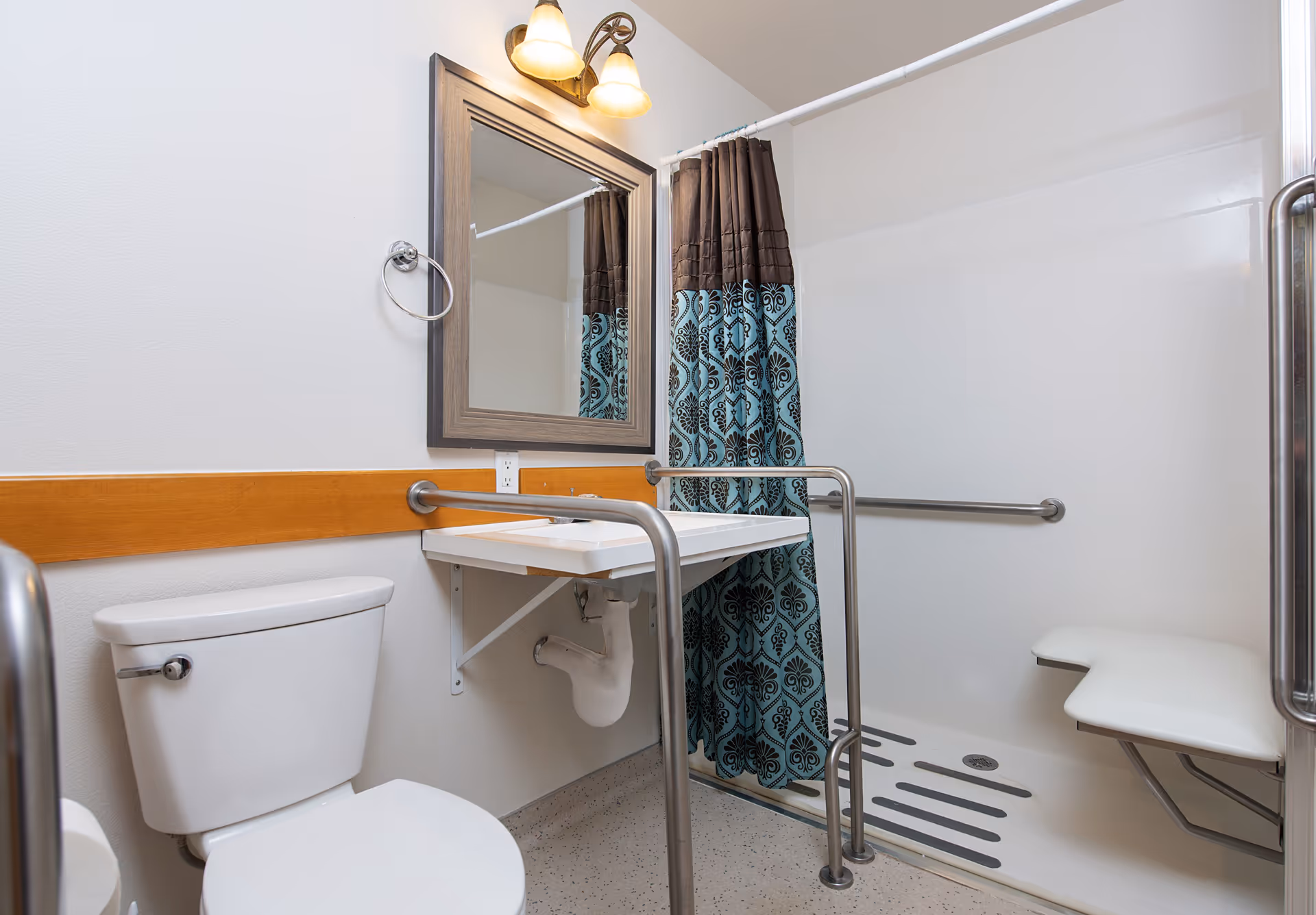 Accessible bathroom with a toilet, a wall-mounted sink with grab bars, a mirror above the sink, and a walk-in shower with a fold-down seat and a patterned shower curtain.