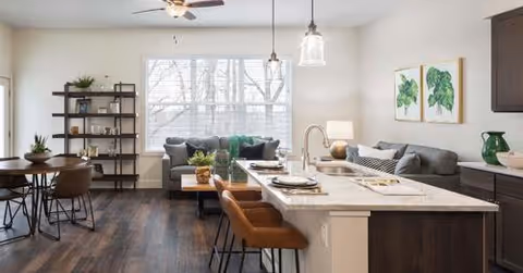 A modern open-concept living area featuring a kitchen island with two brown bar stools, a gray sectional sofa with green and white cushions, a coffee table, and a dining table with four chairs. The room has large windows with white blinds, dark wood flooring, and light-colored walls decorated with two framed leaf prints.