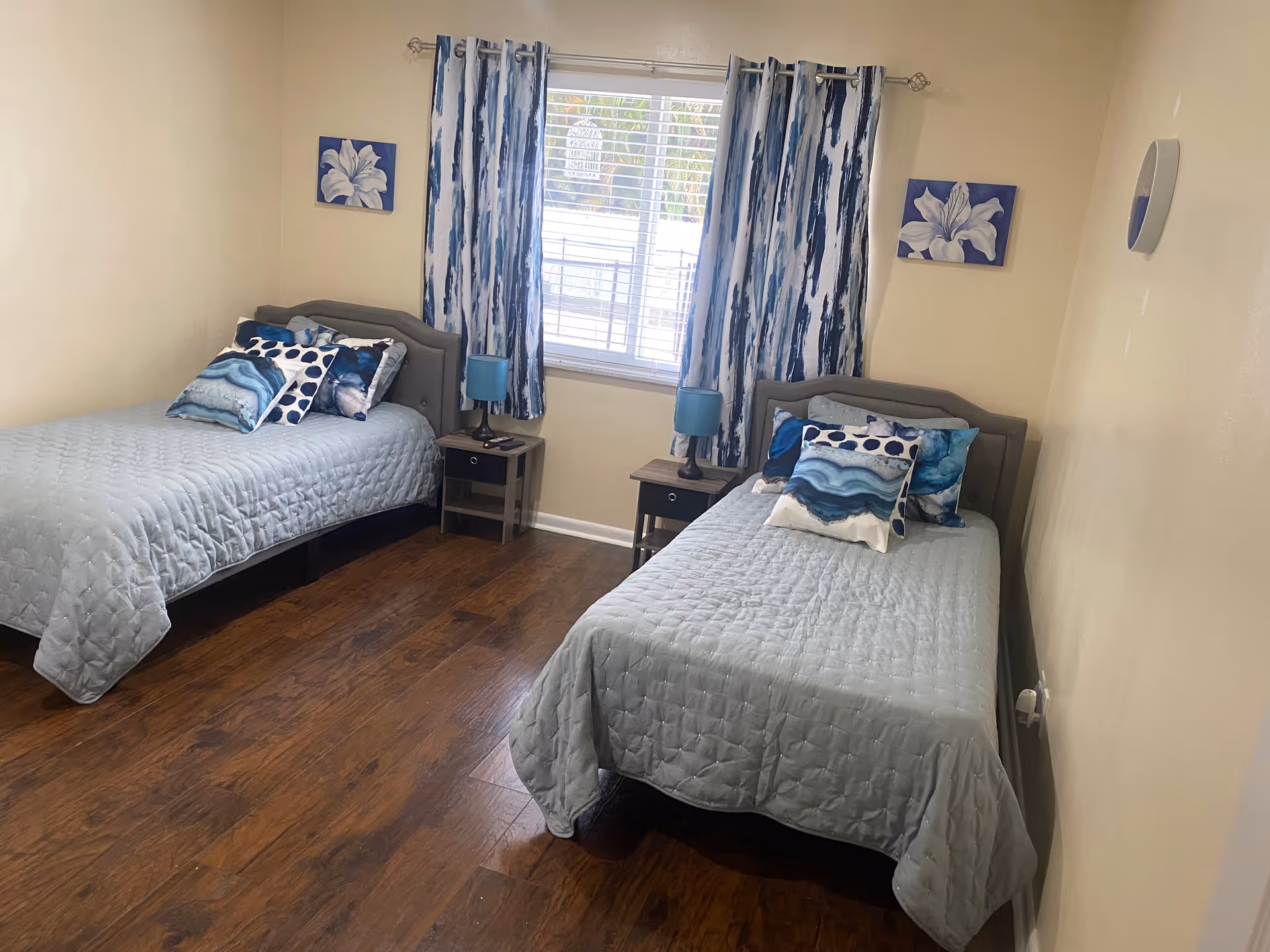 Bright bedroom with two twin beds dressed in blue quilts, nightstands, lamps, and a window with blue-patterned curtains.