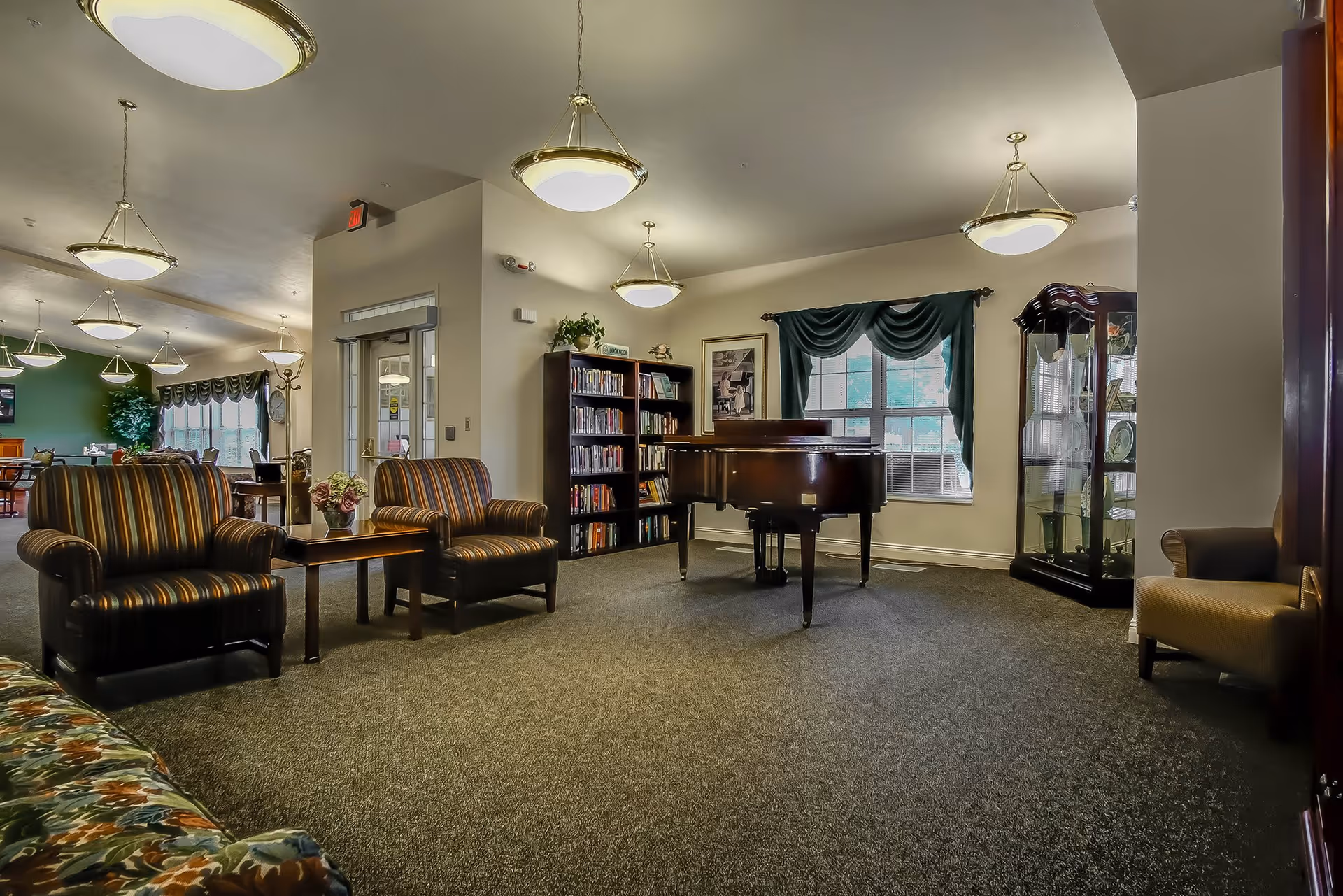 A spacious living room area in a senior living facility featuring a grand piano in the center, two striped armchairs with a small table between them, a bookshelf filled with books, a glass display cabinet, and a window with dark green curtains. The room is well-lit with ceiling lights and has carpeted flooring.