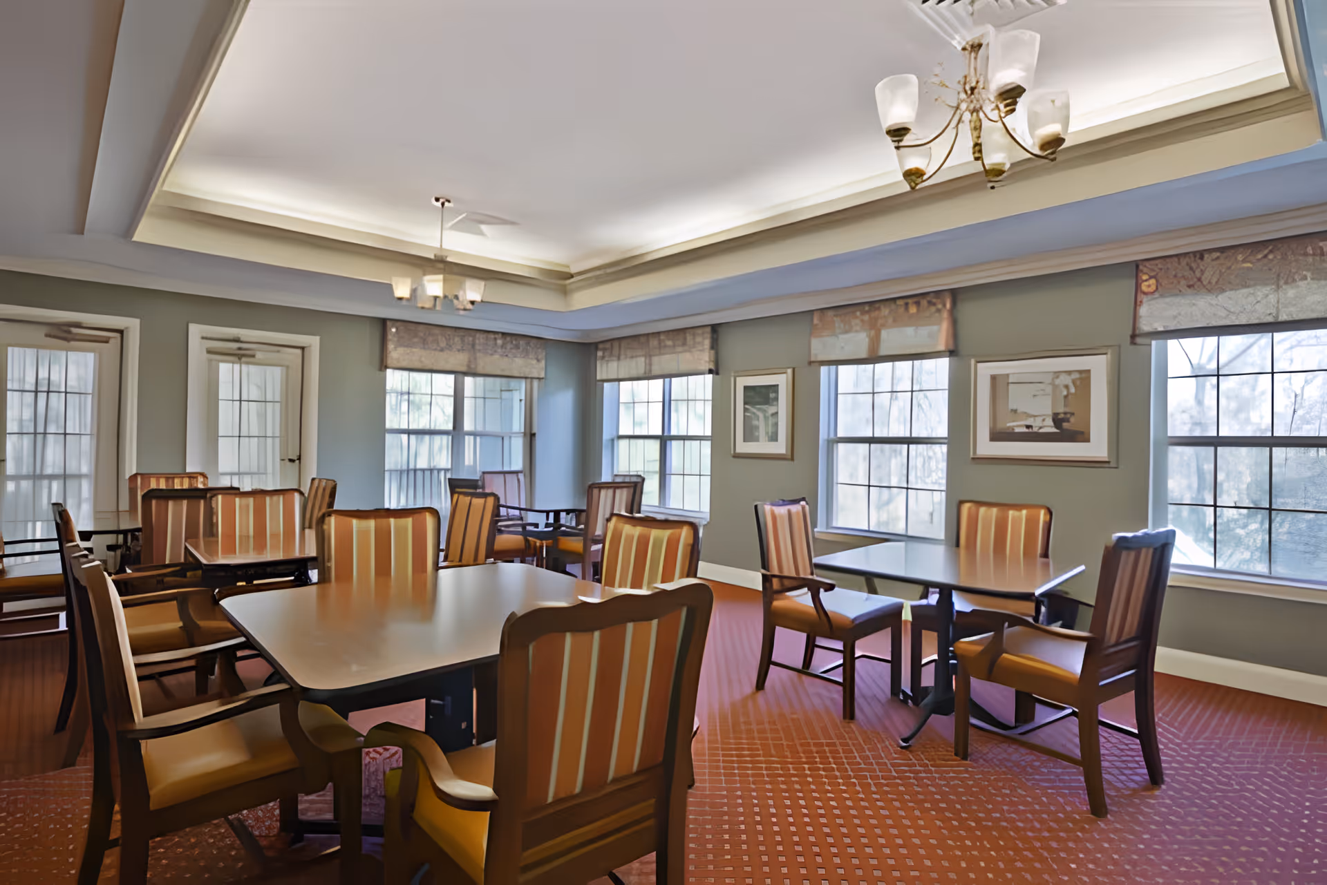 A dining room with multiple square tables and wooden chairs with striped upholstery. The room has large windows with patterned valances, light green walls, a red carpet with a subtle pattern, and two ceiling light fixtures.