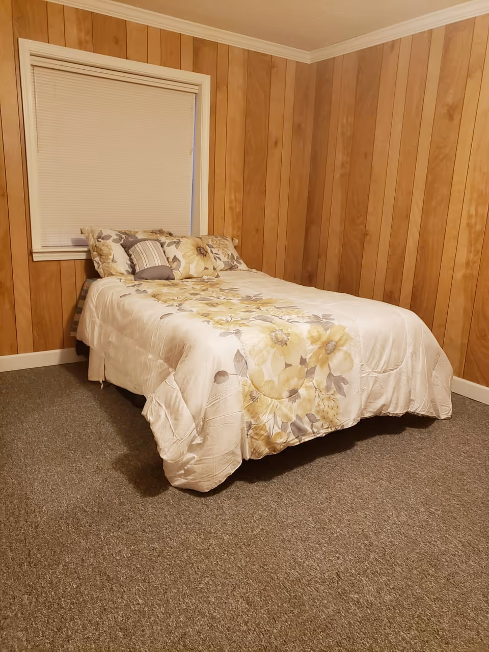 A small bedroom with wood-paneled walls and a window with closed blinds. There is a bed with a floral-patterned comforter and several pillows against the wall.