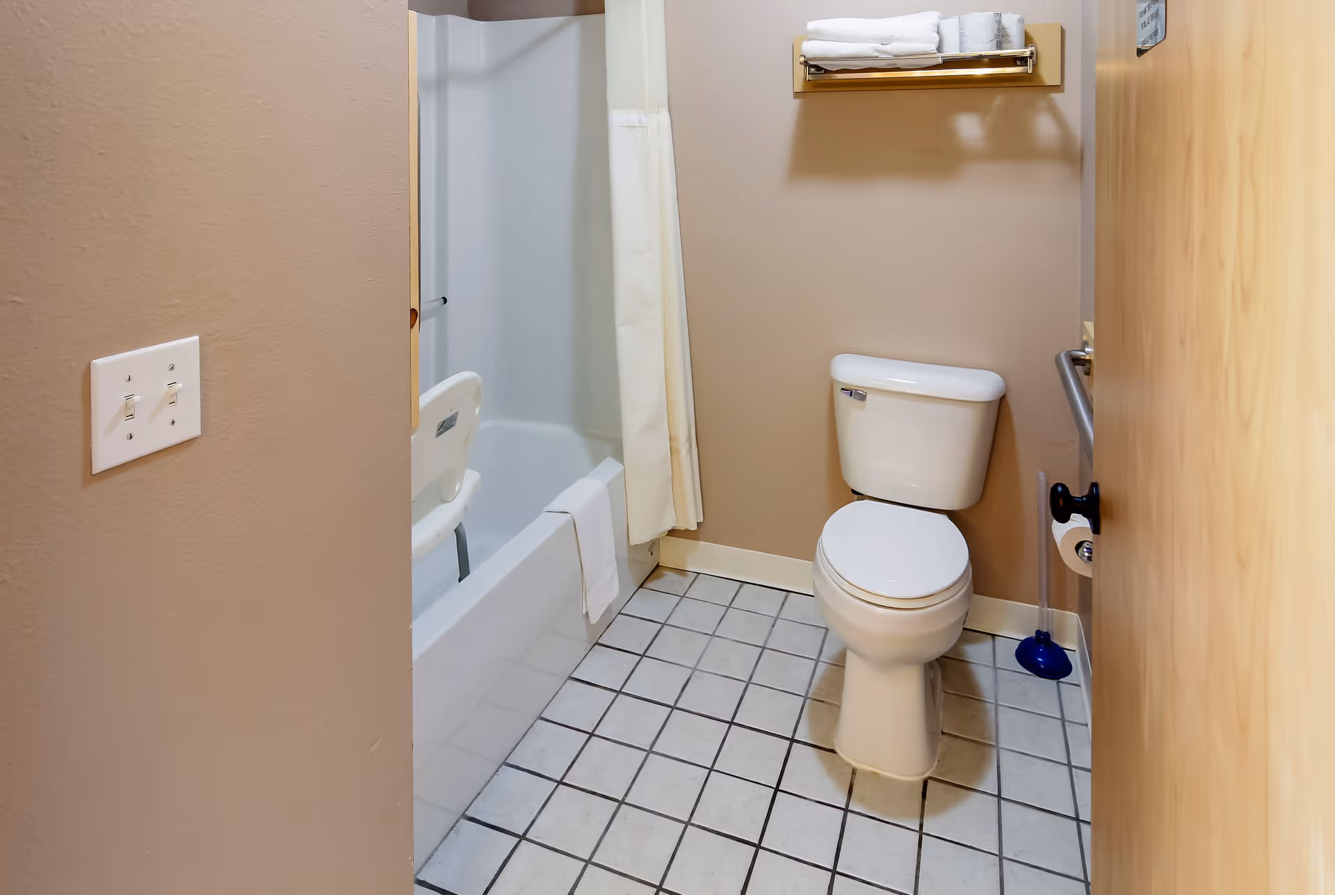 A small bathroom with beige walls and tiled floor. There is a white toilet, a bathtub with a white shower curtain, and a shower chair inside the tub. Above the toilet is a towel rack with folded white towels. A plunger is placed on the floor next to the toilet. A light switch panel is visible on the left wall.