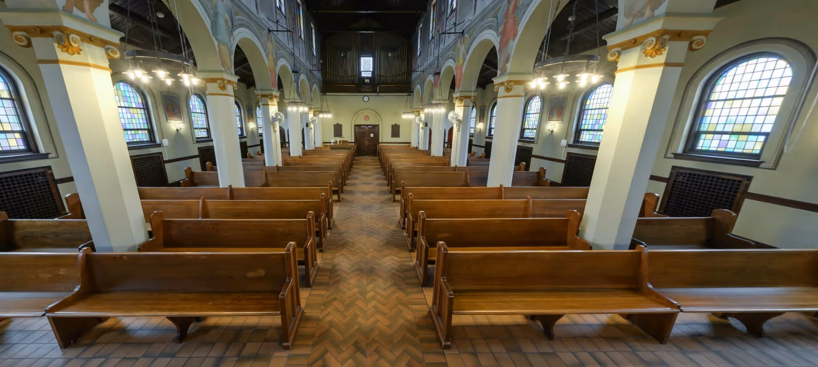 Interior view of a chapel or church with wooden pews arranged in rows on either side of a central aisle. The space features arched columns, stained glass windows, chandeliers, and a high ceiling with visible beams and an organ at the far end.
