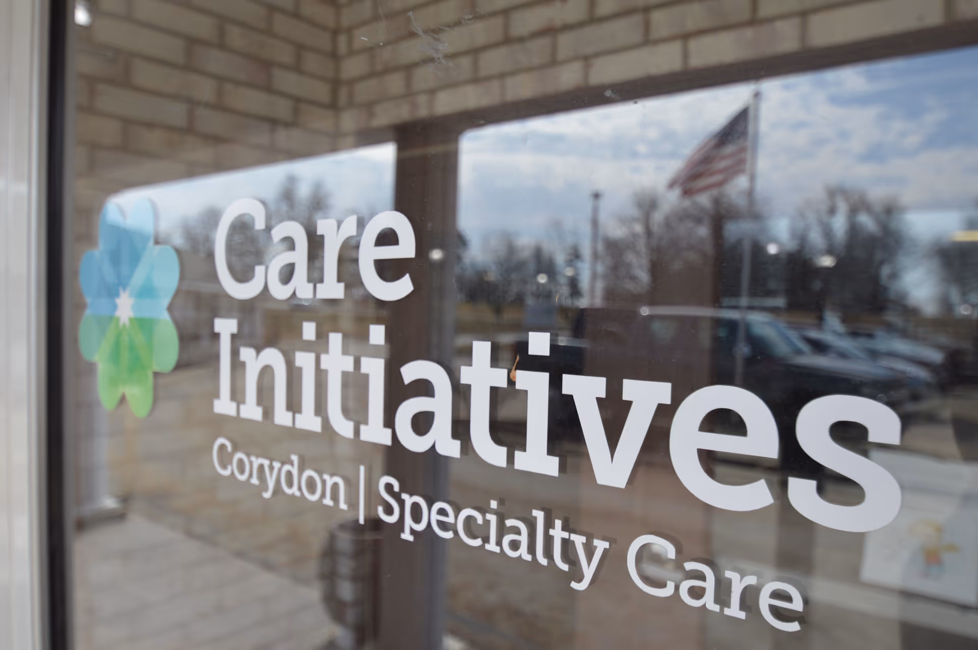 Glass door with the logo and text 'Care Initiatives Corydon Specialty Care' printed on it, with a reflection of an American flag, parked cars, and trees outside.