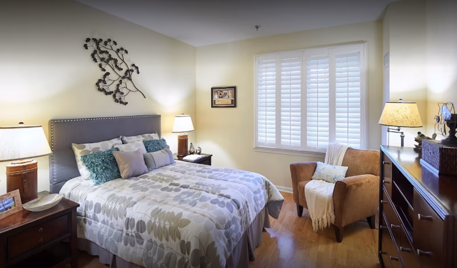 A cozy bedroom with a bed featuring a gray upholstered headboard, patterned bedding, and multiple pillows. There are two bedside tables with lamps on either side of the bed. A brown armchair with a throw blanket and pillow is positioned near a window with white plantation shutters. A dark wooden dresser with a lamp and decorative items is on the right side of the room. The walls are painted light yellow and there is a metal wall art piece above the bed.