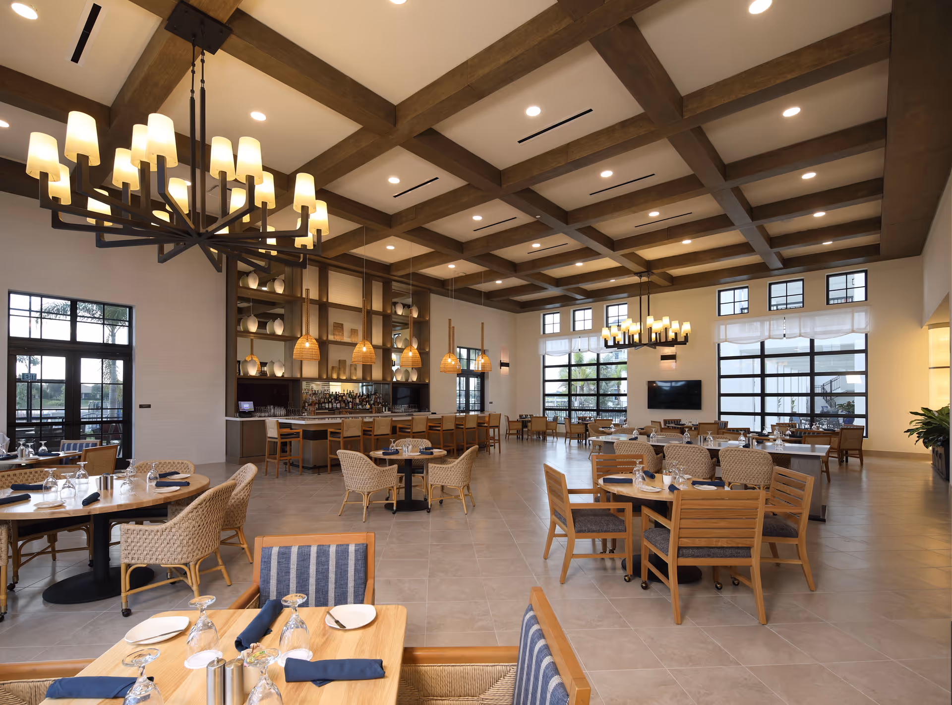 Spacious dining room with set tables and wooden chairs, chandeliers, a bar area, and a coffered ceiling.