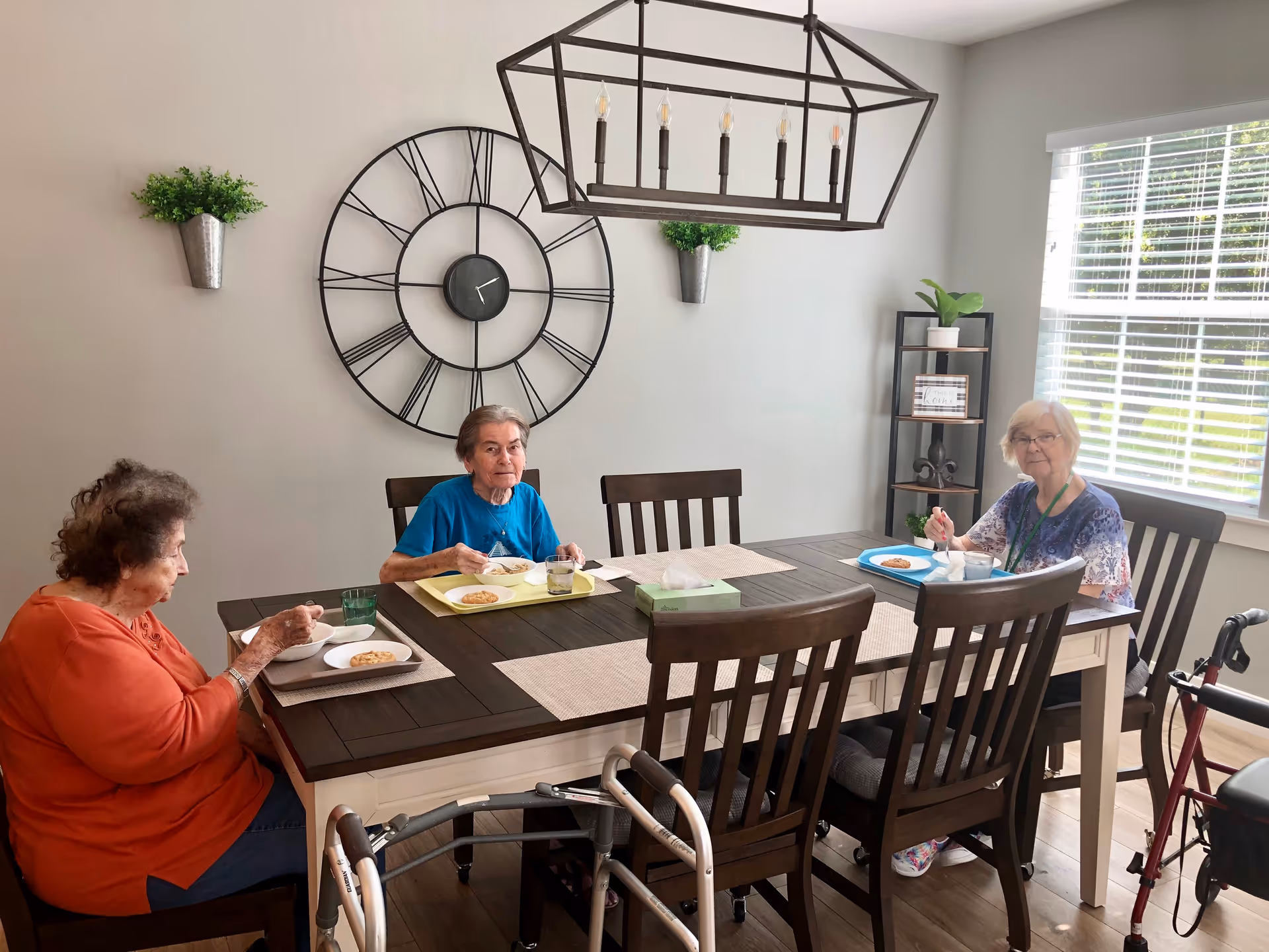 Three elderly women sit around a dining table eating a meal in a bright room with a large wall clock and hanging light fixture.