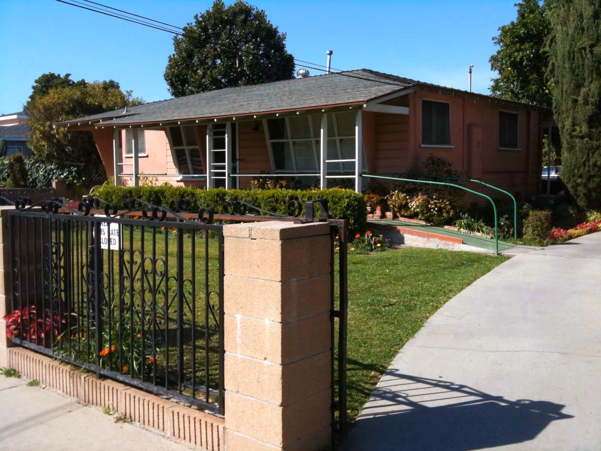 Single-story pink house with a front yard, iron gate, and a green handrail ramp to the entrance.