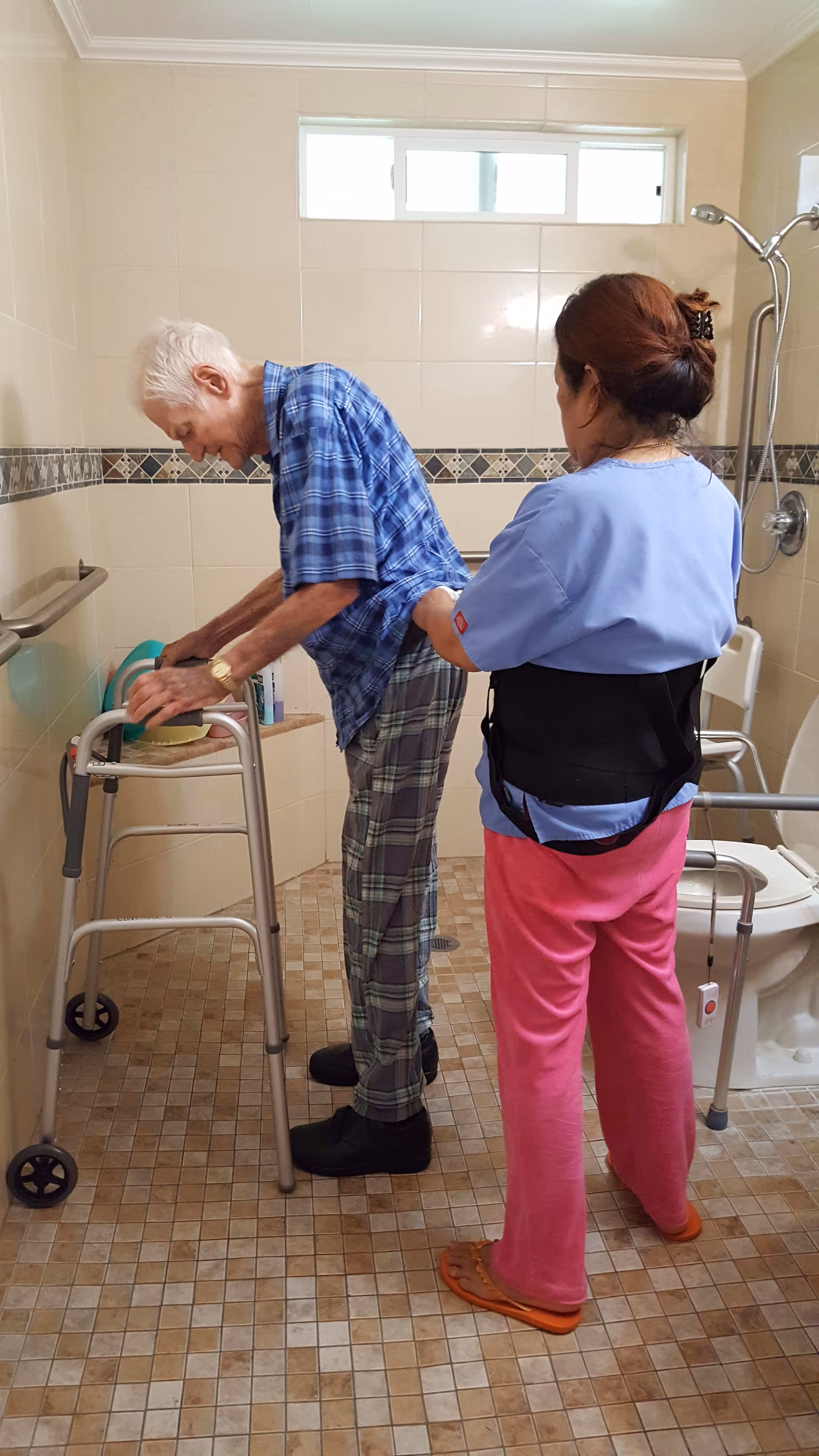 An elderly man using a walker is being assisted by a caregiver in a bathroom. The bathroom has tiled walls and floor, a shower area with a handheld showerhead, and a toilet with grab bars for support.