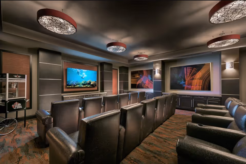 A cozy theater room with two rows of black leather recliner chairs facing a large flat-screen TV mounted on the wall. The room features decorative ceiling lights, abstract artwork on the walls, and a popcorn machine in the corner.