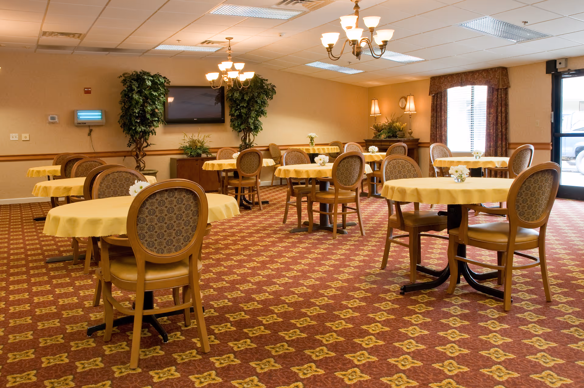 A dining room with multiple round tables covered with yellow tablecloths, each surrounded by wooden chairs with patterned upholstery. The room has a patterned carpet, two large potted plants, a wall-mounted TV, and soft lighting from chandeliers and wall lamps. A window with curtains and a glass door are visible on the right side.