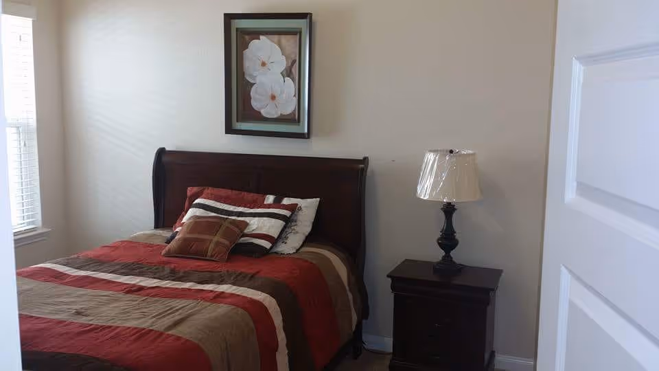 A bedroom with a wooden bed frame and a bed made with a striped red, brown, and beige comforter and several pillows. There is a nightstand with a lamp next to the bed, a window with blinds on the left wall, and a framed picture of white flowers hanging above the bed.